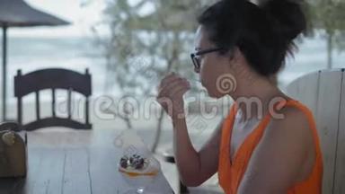 特写镜头，戴眼镜的美女坐在海边的木桌旁的咖啡馆里，吃甜点