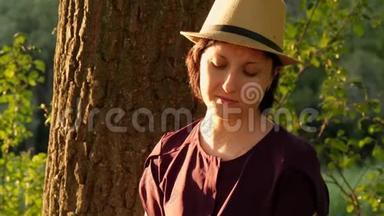 一位戴着帽子的年轻女子坐在公园的一棵树旁，在平板电脑里工作。 务和教育理念..