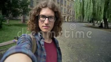 年轻英俊的博客写手戴着卷发眼镜拍摄视频或自拍，站在大学附近的街道上