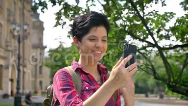 一位留着短发、通过电话和微笑视频<strong>聊天</strong>的快乐年轻女子站在大学附近的公园里，<strong>学生</strong>