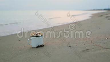一个白色的<strong>金属</strong>杯，里面<strong>装满</strong>了贝壳，矗立在沙质的海岸上，面对着模糊的海浪