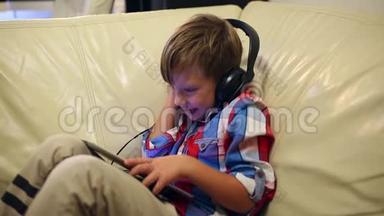 快乐的小男孩戴着耳机在平板电脑上播放音乐，坐在家里的沙发上听音乐