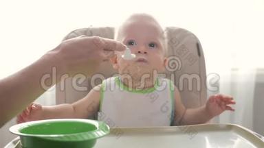 婴儿坐在孩子们`小桌子旁。 妈妈给孩子喂粥。 妈妈用勺子给婴儿食物。