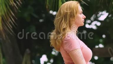一个美丽的金发女人，在一个刮风的日子里站在棕榈树上，她看着镜头微笑