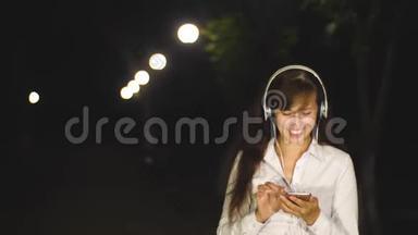 夜晚，女孩在城市公园里走着灯笼灯。 戴着耳机听音乐的漂亮女孩