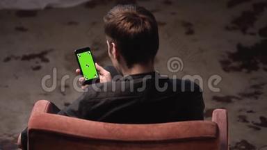 在黑暗的房间里，棕色头发的年轻人坐在椅子上敲着iPhone绿色屏幕。 库存数据