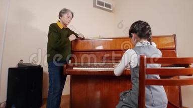 音乐课。 女孩弹钢琴，年长的老师站在钢琴附近，帮助弹钢琴。 斜视图