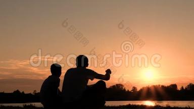 两个人在河岸或湖边交流的剪影。 父子俩坐在水边看着