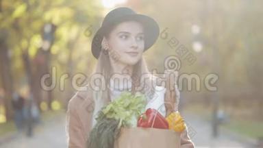 年轻漂亮的女人穿着时髦的外套在公园里散步，拿着一包产品，微笑着环顾四周。