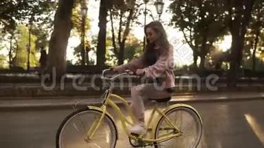 在夏季城市公园的街道或林荫大道上骑自行车的头发很长的女孩。 穿着粉色休闲服