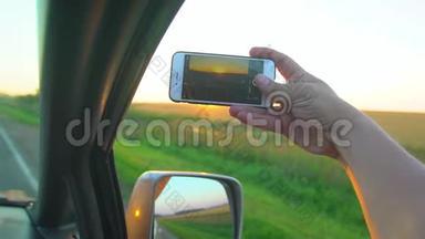 男人手在日落时在手机，智能手机上拍照。 一个人在阳光下从车窗`手`阳光照射着