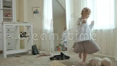 穿着粉红色裙子扎着辫子的开朗女孩试着穿着成年妈妈`衣服、连衣裙、高跟鞋和在前面跳舞
