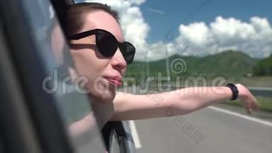 戴着太阳镜的快乐女孩斜倚在车窗外，享受着在山里的旅行。 年轻女子望着窗外
