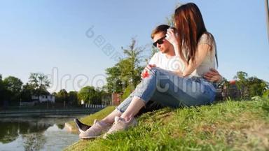 浪漫的夫妻喝咖啡，笑着坐在池塘附近的绿草上。 夫妻信任，爱情和幸福的概念