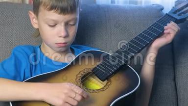 男孩坐在沙发上弹吉他。 学习演奏乐器的概念..