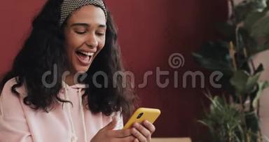 年轻迷人的女孩在智能手机上阅读好消息，坐在床上玩得很开心。 她唱歌，跳舞，快乐