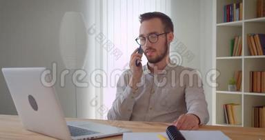 一位年轻英俊的白种人商人坐在笔记本电脑前打电话的特写镜头。