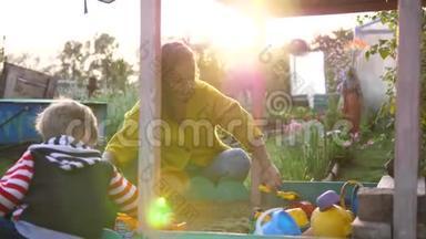 一位年轻的母亲，带着一个孩子在沙箱里玩耍。 夏日，操场上的阳光