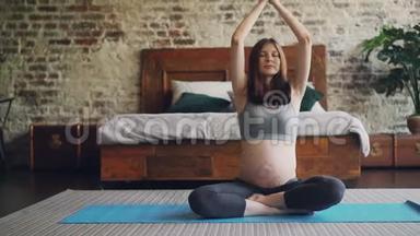 漂亮的孕妇正在做瑜伽，坐在莲花的位置，卧室的地板上，明亮的垫子上，女孩在举手