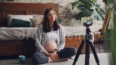 快乐的怀孕女孩正在用三脚架上的智能手机为她的在线视频制作视频，女孩正在和订户交谈。