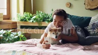 快乐的<strong>狗主人</strong>正在和可爱的宠物一起躺在床上，抱着智能手机，摆姿势，拥抱动物。