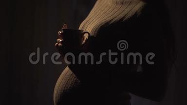 不明身份的怀孕女孩站在一个温暖的毛衣在黑暗的房间里，晚上拿着一杯热茶。 夫人