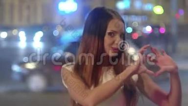 夜晚城市里美丽的年轻女子在背景上用模糊的交通<strong>灯</strong>照相机显示出心脏形状的<strong>手指</strong>