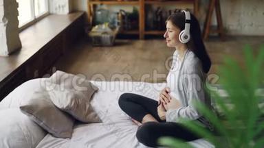 快乐的期待妈妈抚摸她的肚子，通过耳机听音乐，躺在舒适的双人床上