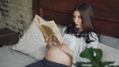 微笑的女孩期待着母亲，大声朗读书给她未出生的婴儿，微笑着躺在现代公寓的床上