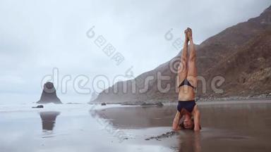 在海洋边，一个女人在瑜伽倒立和冥想。 健康的生活方式。 站在沙滩上平衡你的头