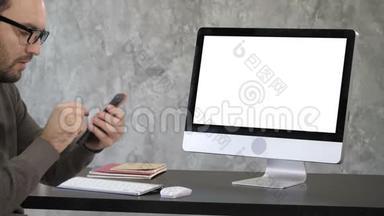 迷人的胡须男检查他的牙齿在他的手机自拍相机附近的电脑显示器。 白色显示器。
