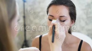 一位年轻的金发碧眼的化妆师用一个<strong>遮瑕</strong>膏在她脸上画一个年轻的黑发女孩