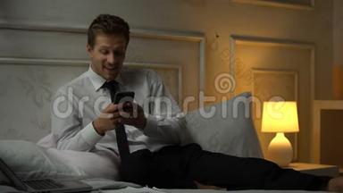 微笑的男人穿着西装，坐在床上，在智能手机、交友网站、应用程序上发短信