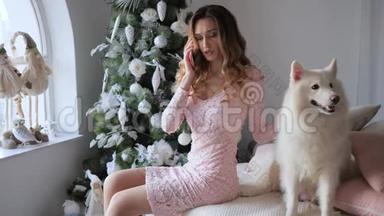 糟糕的圣诞节，沮丧的女孩坐在靠近白狗的床上，在圣诞树的背景下用手机交谈