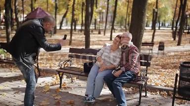一个成年的儿子和年迈的父母在秋天公园散步，给坐在长凳上的父母拍照。