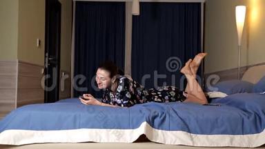 美丽的黑发女孩躺在床上用<strong>手机</strong>。 网上<strong>约会</strong>