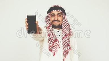 穿着民族服装微笑的阿拉伯人展示了智能手机和广泛的微笑。 一个男人展示一部现代手机。 商业和办公室。 阿拉伯
