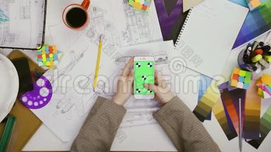 双手顶视。 女设计师使用她的智能手机，桌子上有绿色屏幕，<strong>滚动</strong>新闻，<strong>照片</strong>