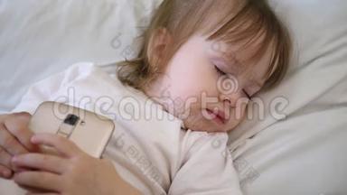 可爱的婴儿睡在床上智能手机。 孩子躺在枕头上拿着一块药片