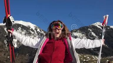 弯曲的滑雪者在山上的背景下在雪中欢乐。 戴镜镜眼镜的女人，手里拿着滑雪服和滑雪板