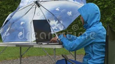 穿防水衣服的商务女士在雨中的野餐桌上工作笔记本电脑