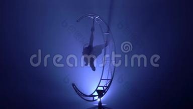 体操运动员倒挂在旋转结构的月亮上的一根绳子上。 蓝色烟雾背景。 慢<strong>动作</strong>。 <strong>剪影</strong>
