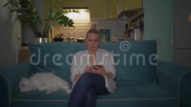 一个年轻的女人晚上和一只猫坐在沙发上用电话。 4K