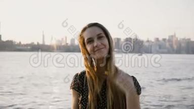 快乐微笑的白人女孩在镜头前摆姿势的肖像，在纽约日落海滩天际线4K戴上太阳镜。