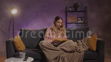 一个年轻的女人坐在沙发上看书。 女孩在温暖的房间里的舒适沙发上做梦