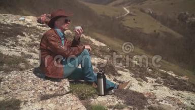 山上坐着一个男人，戴着牛仔帽、皮夹克、蓝色牛仔裤和眼镜。 一个人喝热水瓶里的茶。