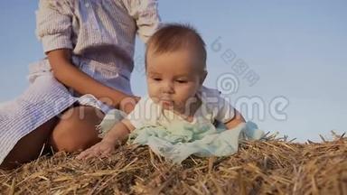 可爱的孩子穿着白色衬衫，在夏天的干草堆上玩着一件蓝色裙子的年轻母亲。 女人抱着孩子玩