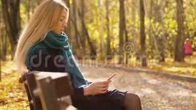 年轻漂亮的金发女人坐在长凳上，在秋日公园使用智能手机。 3840x2160