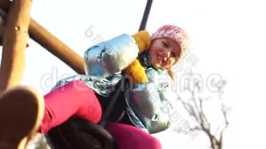 可爱的女孩在美丽的冬日在秋千上玩得很开心。 这个女孩穿着一顶粉红色的针织帽子和一顶蓝色发亮的帽子