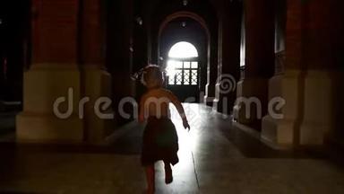 一个穿着连衣裙的小女孩正沿着黑暗的走廊在柱子上奔跑。 玩追赶。